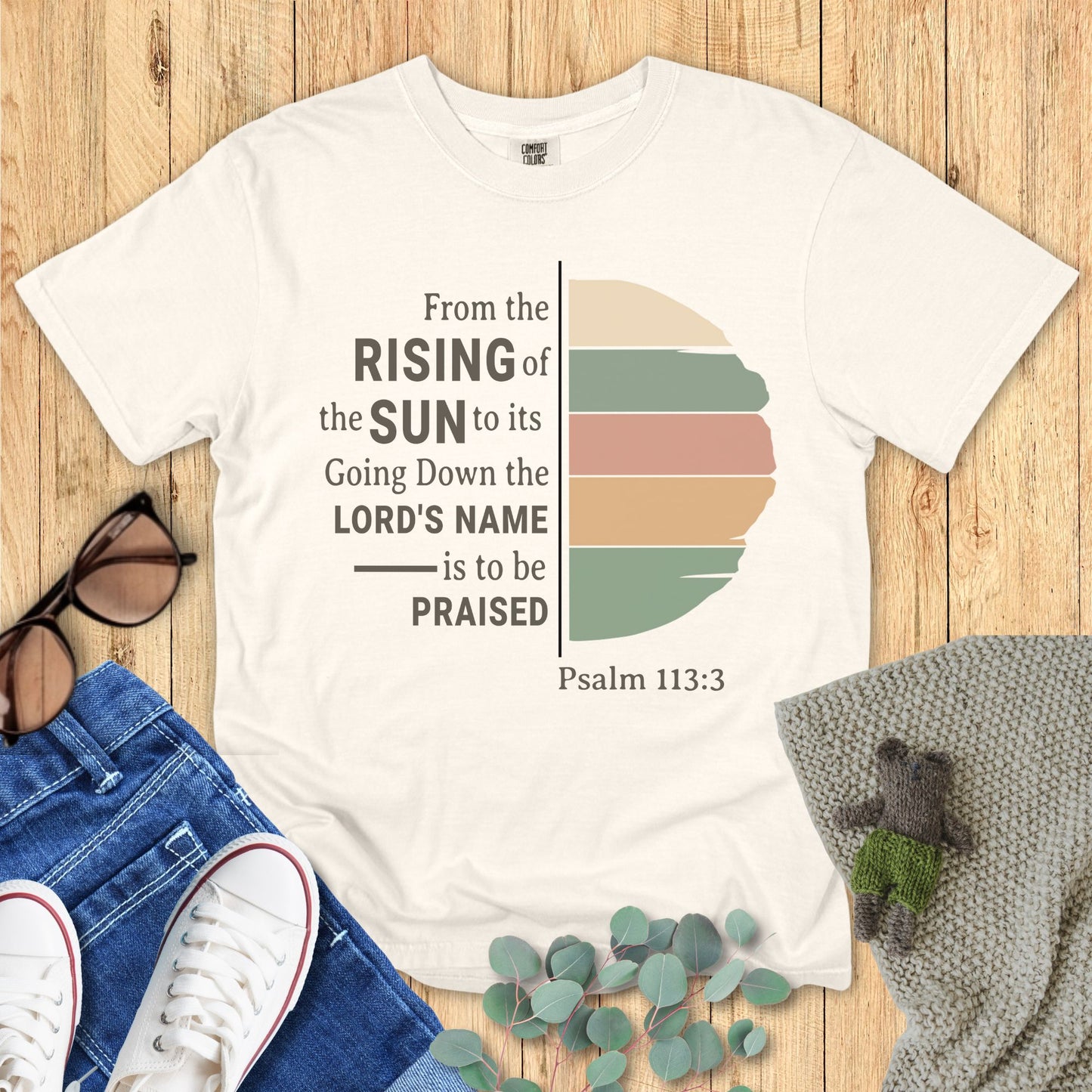 Ivory White t-shirt with sunset design and text on a wooden surface with accessories.