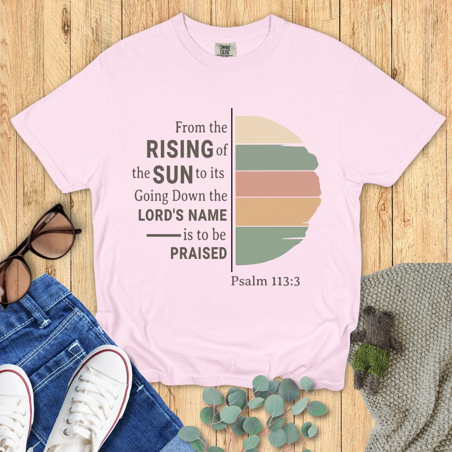 Pink t-shirt with sunset design and Psalm 113:3 text on a wooden surface with jeans and sneakers.