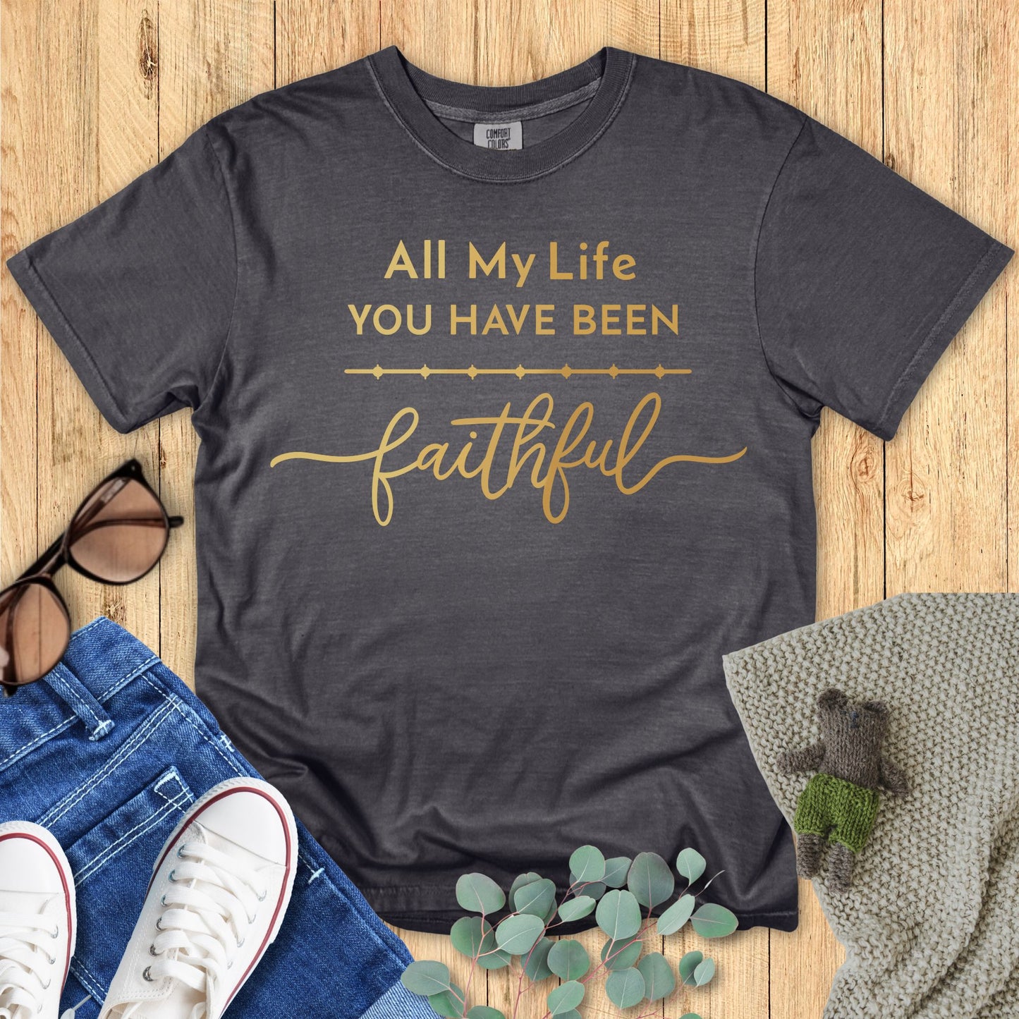 Gray t-shirt with gold text on a wooden surface with accessories.