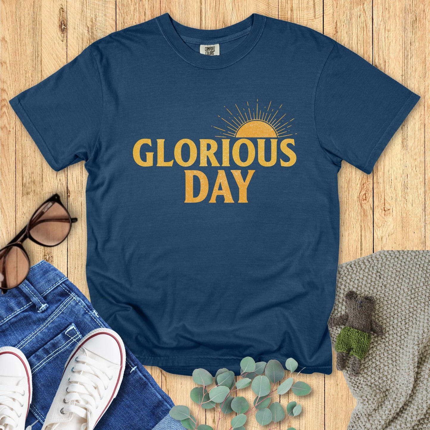 Blue t-shirt with 'Glorious Day' text on a wooden surface with accessories.