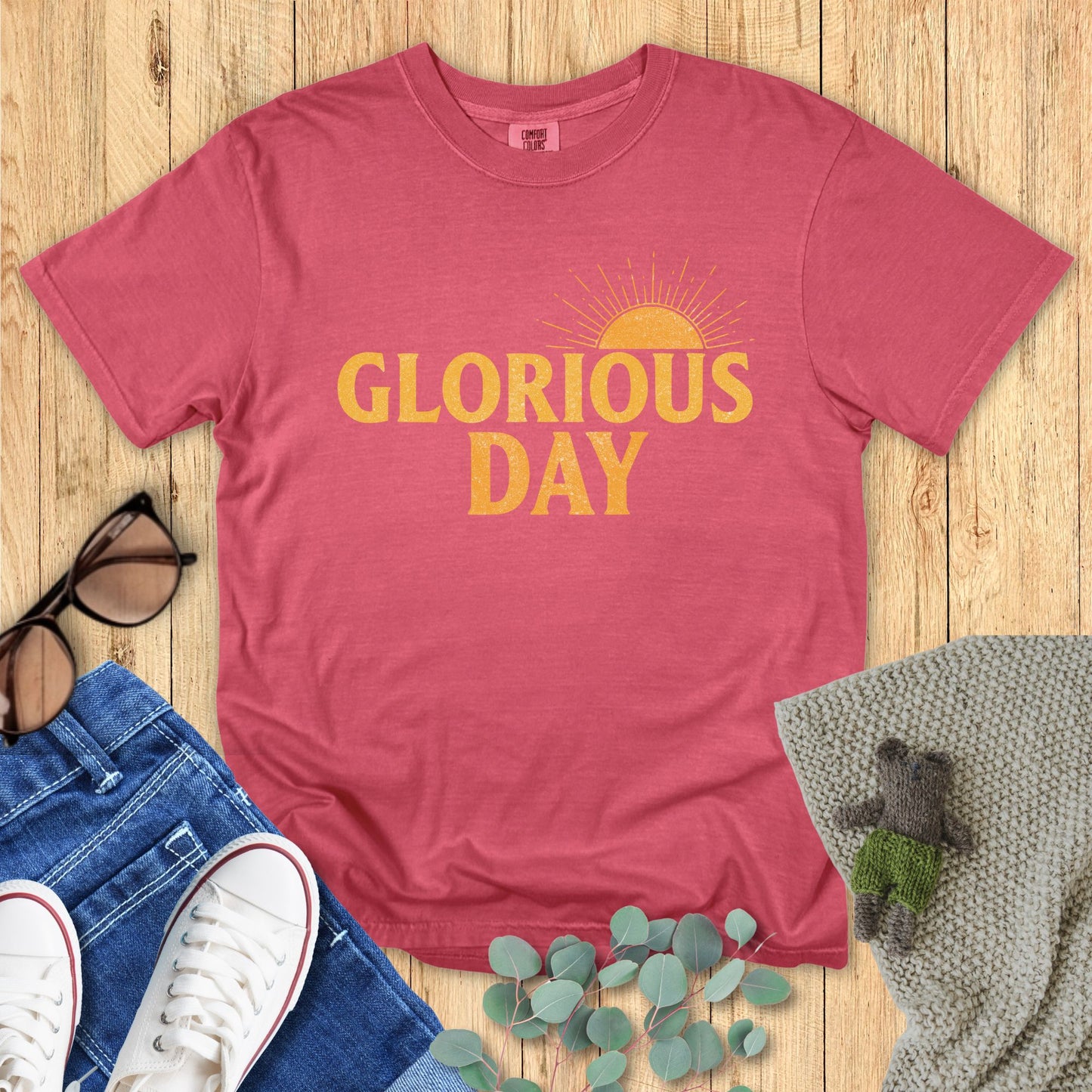 Crimson Red t-shirt with 'Glorious Day' text on a wooden surface with accessories.