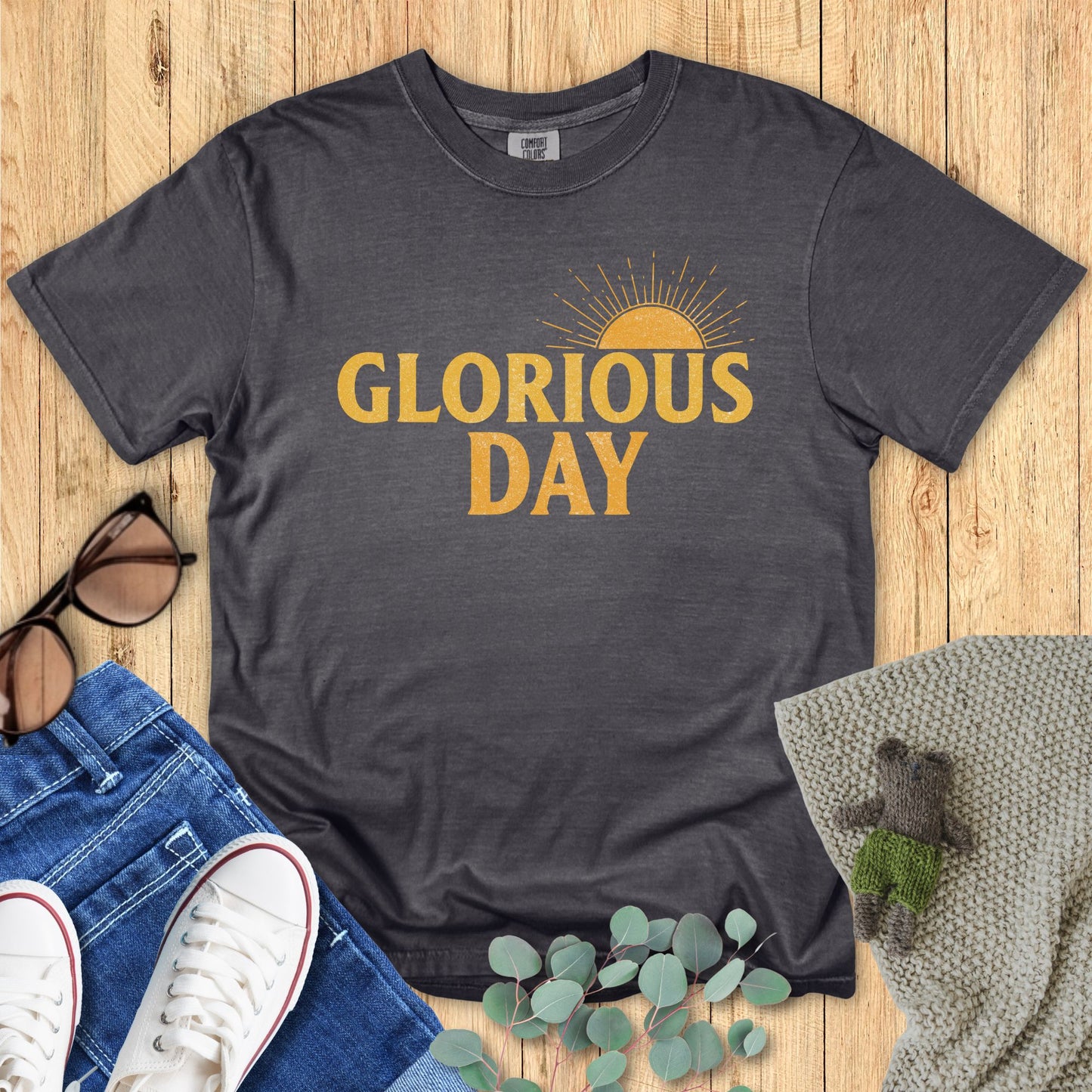 Gray t-shirt with 'Glorious Day' text on a wooden surface with accessories.