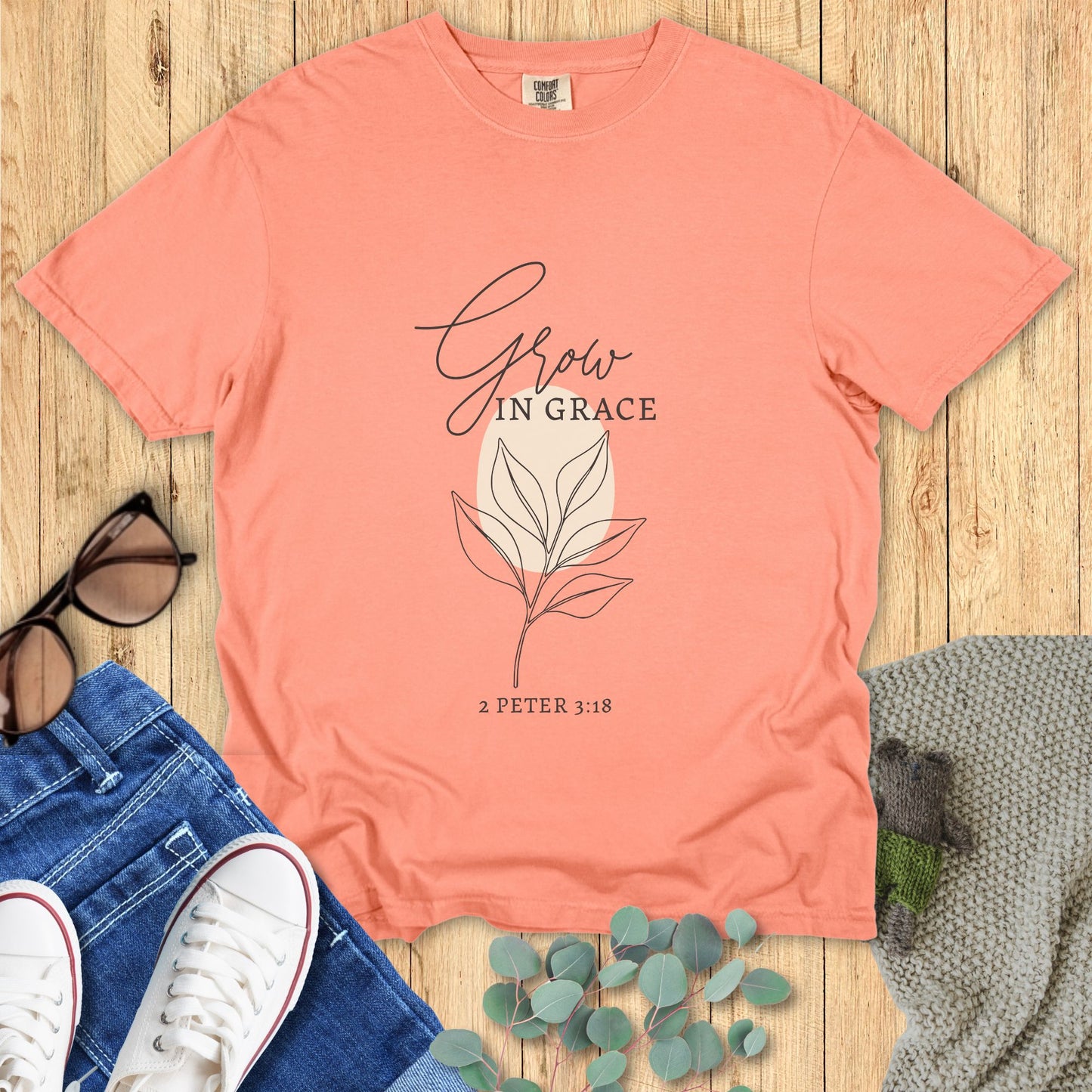 Coral t-shirt with 'Grow in Grace' text and leaf design on a wooden surface with accessories.