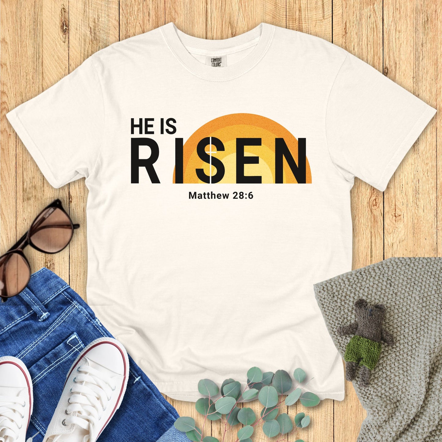 Ivory White t-shirt with 'HE IS RISEN' text on a wooden surface with accessories.
