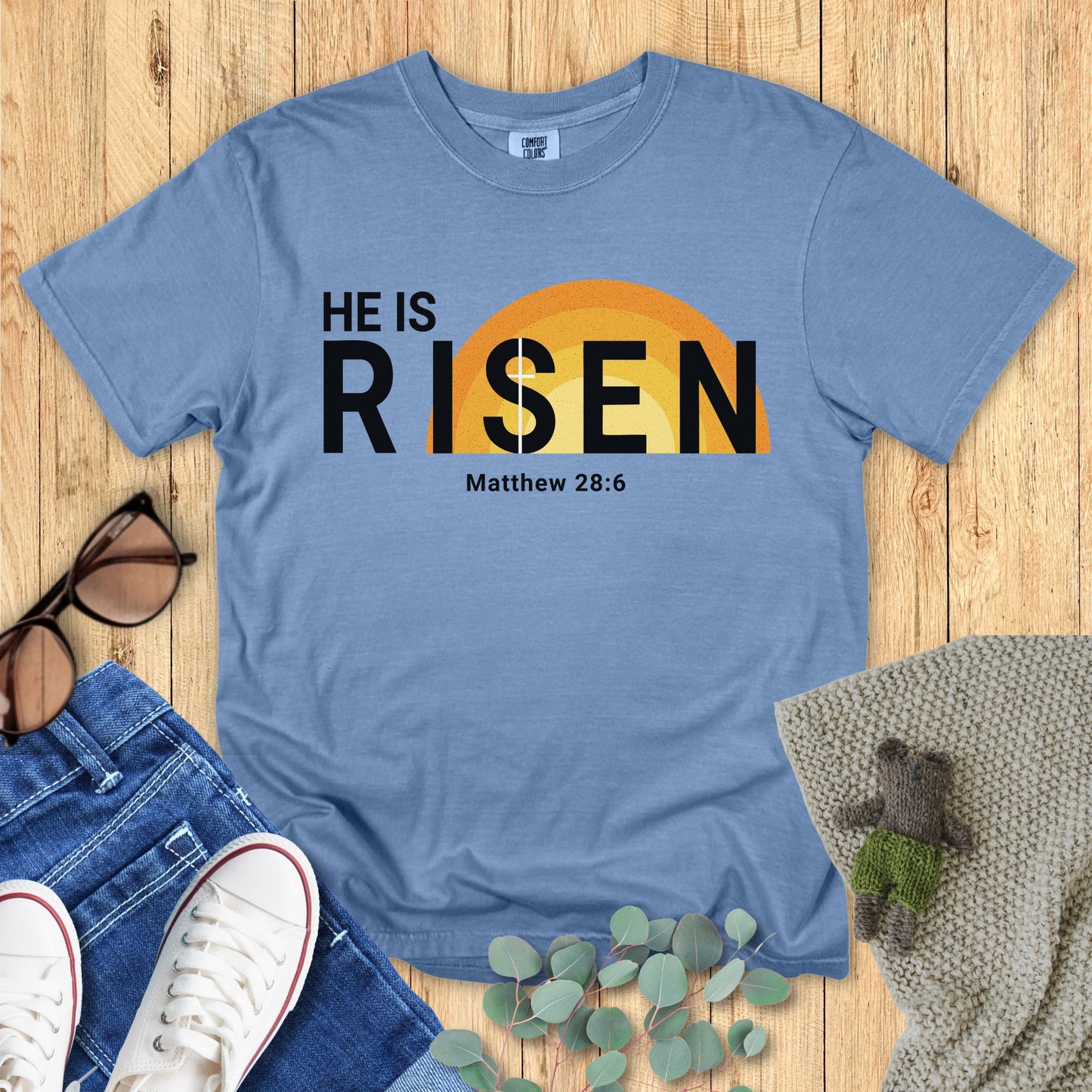 Blue t-shirt with 'HE IS RISEN' text on a wooden surface with accessories.