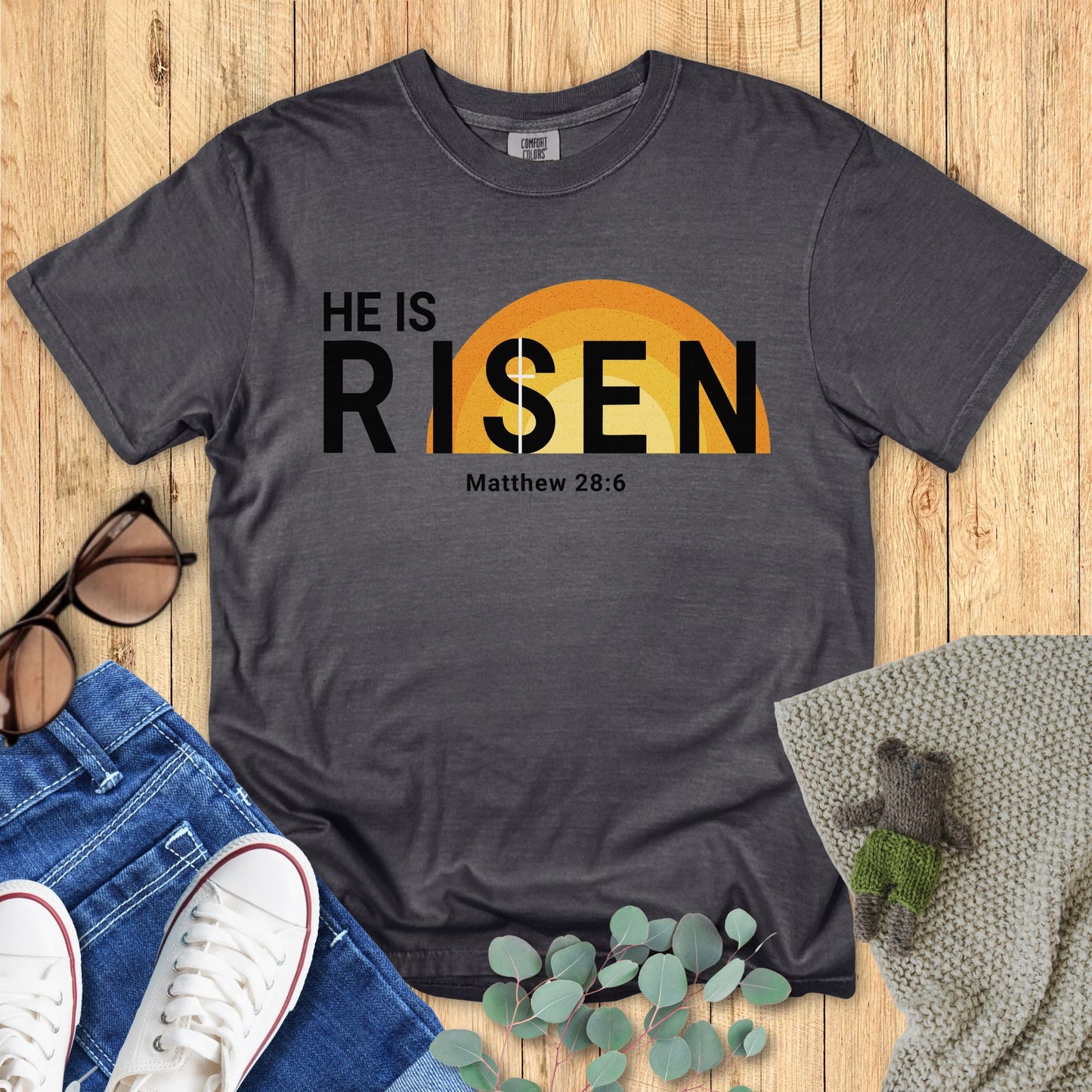 Gray t-shirt with 'HE IS RISEN' design on a wooden surface with accessories.