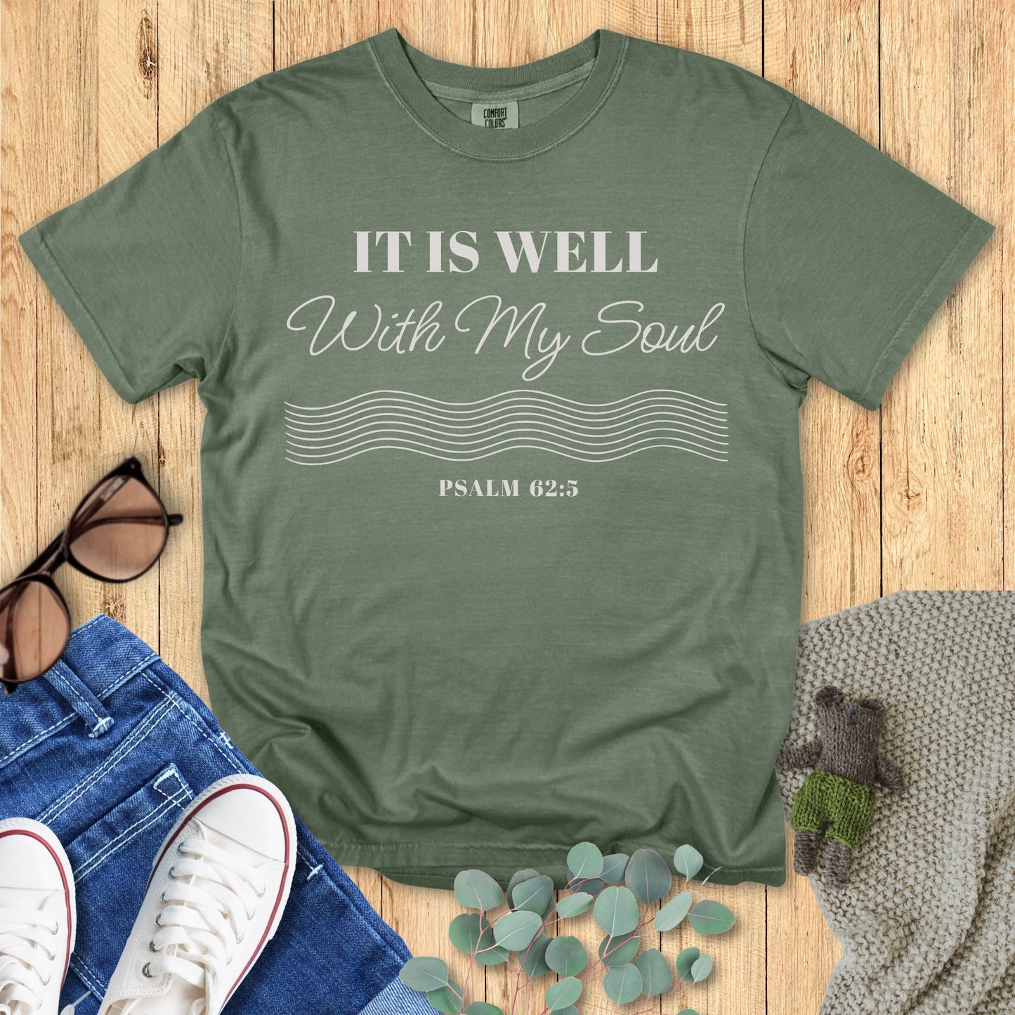 Green t-shirt with 'It is Well with My Soul' text on a wooden surface with accessories.