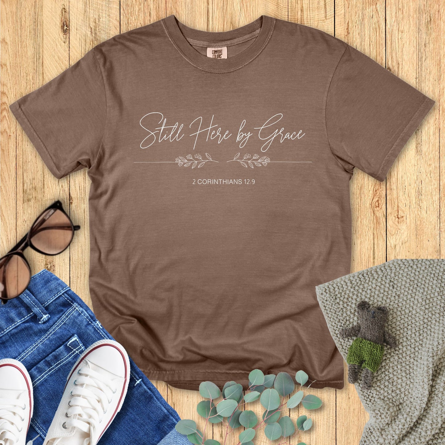 Brown t-shirt with 'Still Here by Grace' text on a wooden surface with accessories.