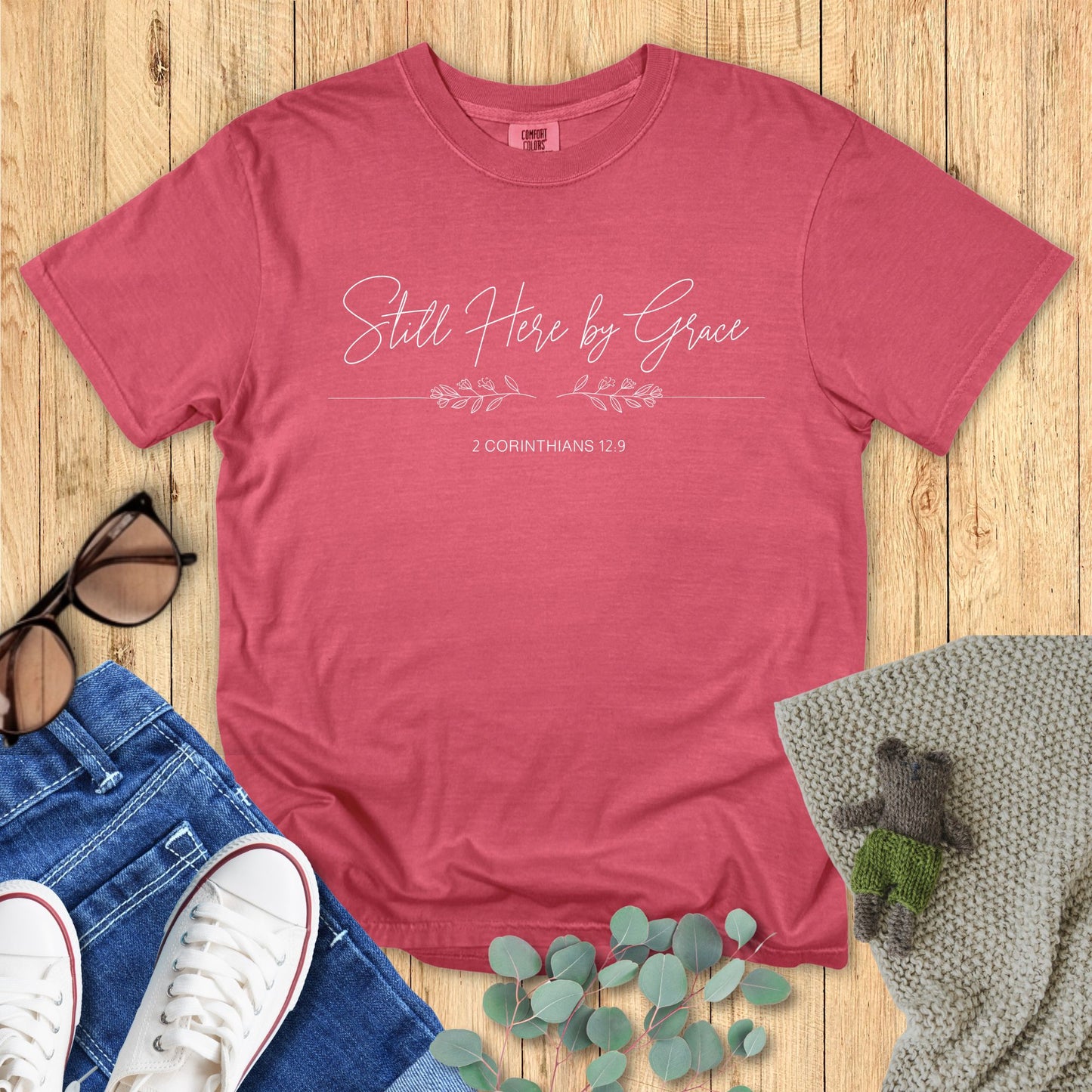 Red t-shirt with 'Still Here by Grace' text on a wooden surface with accessories.