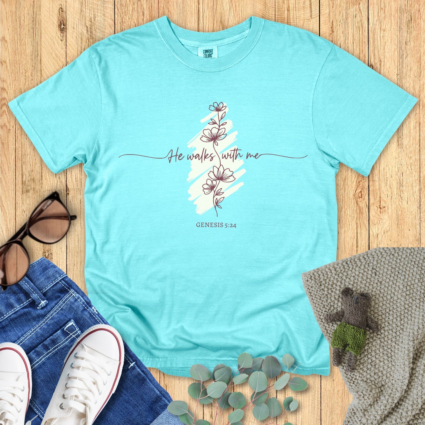 Light blue t-shirt with floral design and text on a wooden surface with accessories.