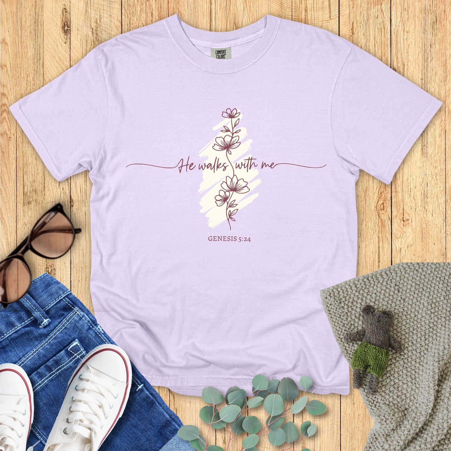 Lavender t-shirt with floral design and text on a wooden surface with accessories.