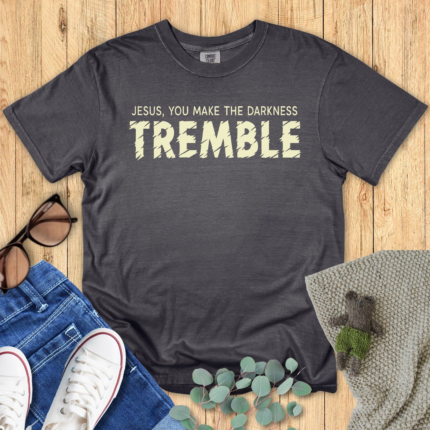Dark gray t-shirt with 'Jesus, you make the darkness tremble' text on a wooden surface with accessories.