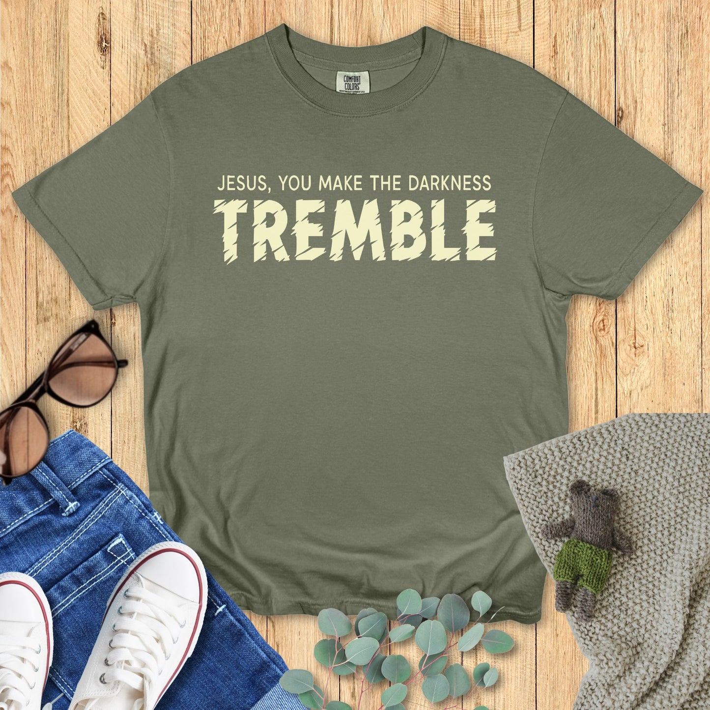 Green t-shirt with 'Jesus, you make the darkness tremble' text on a wooden surface with jeans and shoes.
