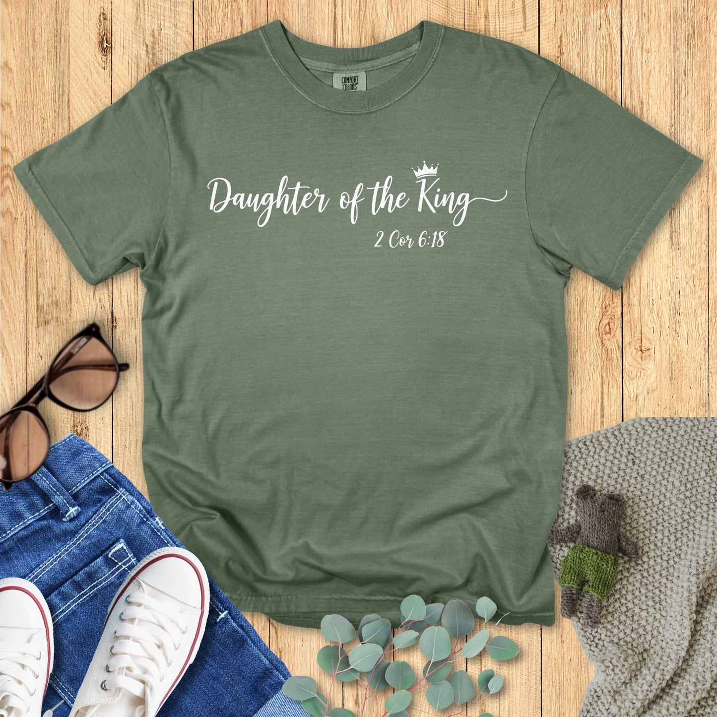 Green t-shirt with 'Daughter of the King' text on a wooden surface with accessories.