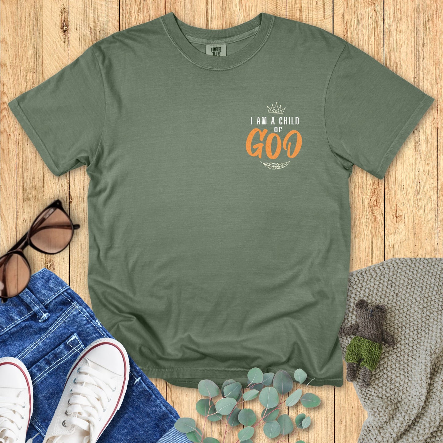 Green t-shirt with 'I am a Child of Goo' text on a wooden surface with accessories.