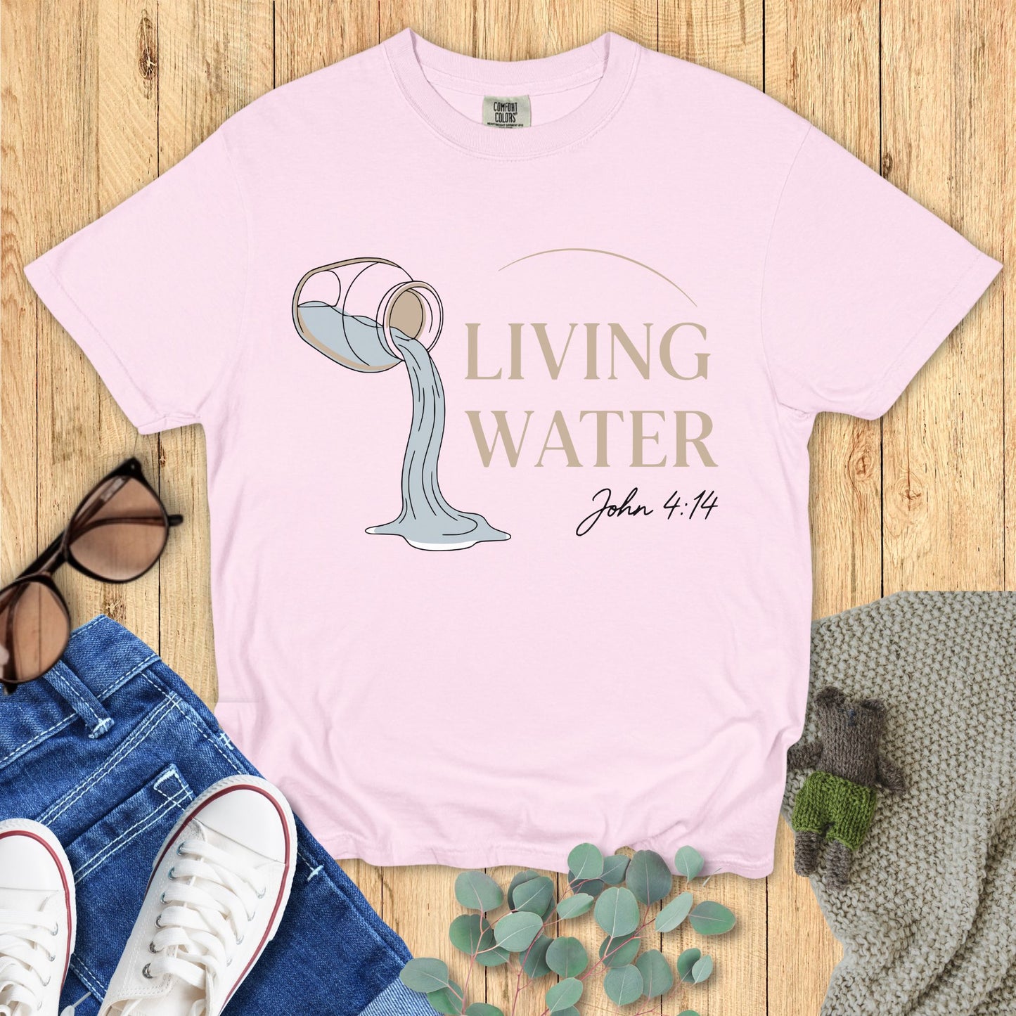 Pink t-shirt with 'Living Water' design and text on a wooden surface with accessories.