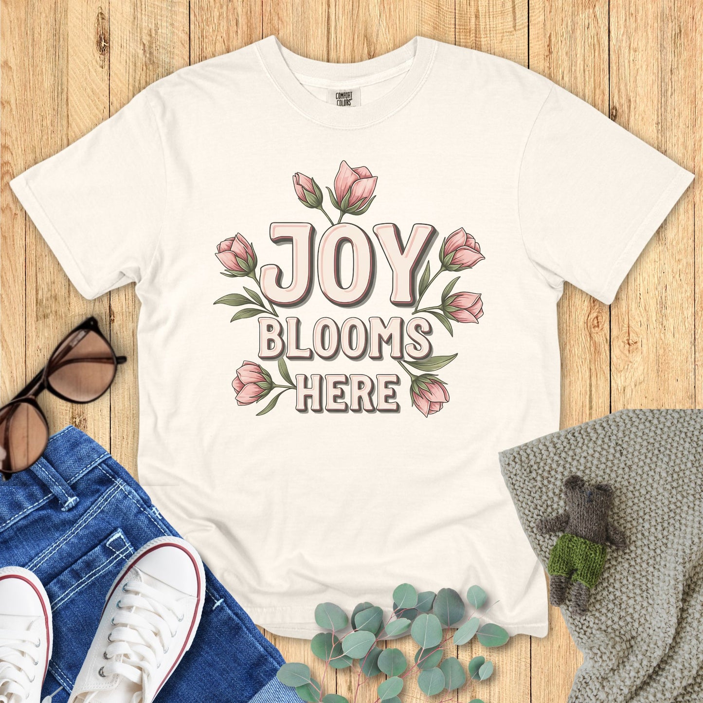 Ivory White t-shirt with 'Joy Blooms Here' design on a wooden surface with accessories.