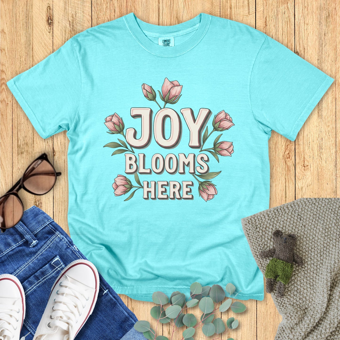 Light blue t-shirt with 'Joy Blooms Here' design on a wooden surface with accessories.