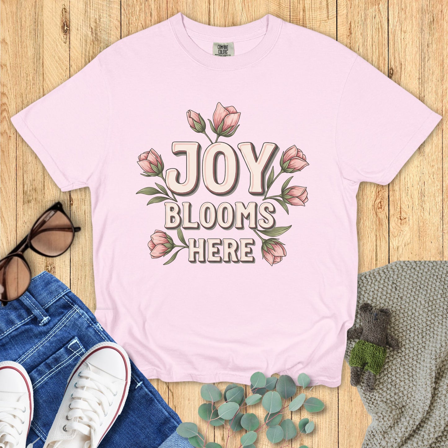 Pink t-shirt with 'Joy Blooms Here' design on a wooden surface with jeans and sneakers.