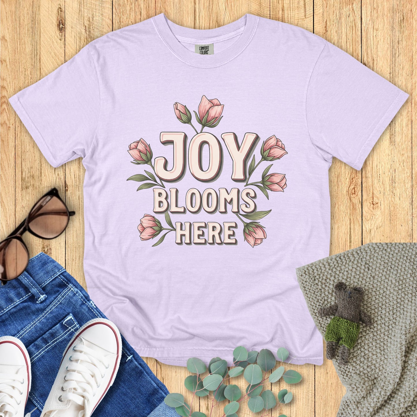 Lavender t-shirt with 'Joy Blooms Here' design on a wooden surface with accessories.