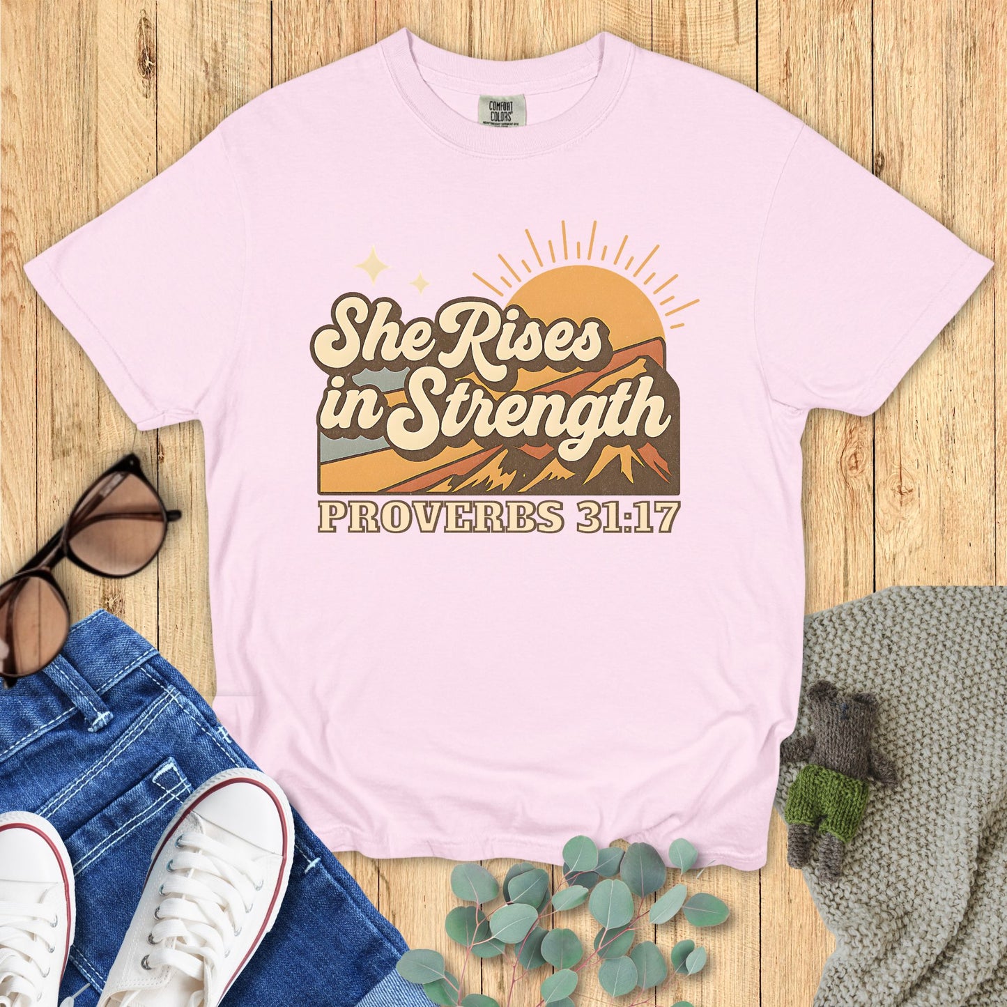 Pink t-shirt with 'She Rises in Strength Proverbs 31:17' design on a wooden surface with jeans and sneakers.