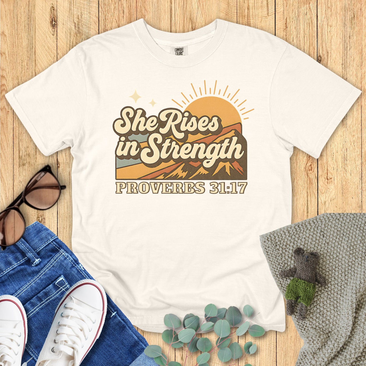 Ivory White t-shirt with 'She Rises in Strength' design on a wooden surface with jeans, sneakers, and a plant.