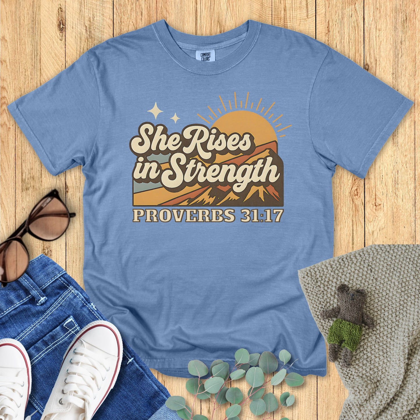 Blue t-shirt with 'She Rises in Strength Proverbs 31:17' design on a wooden surface with accessories.