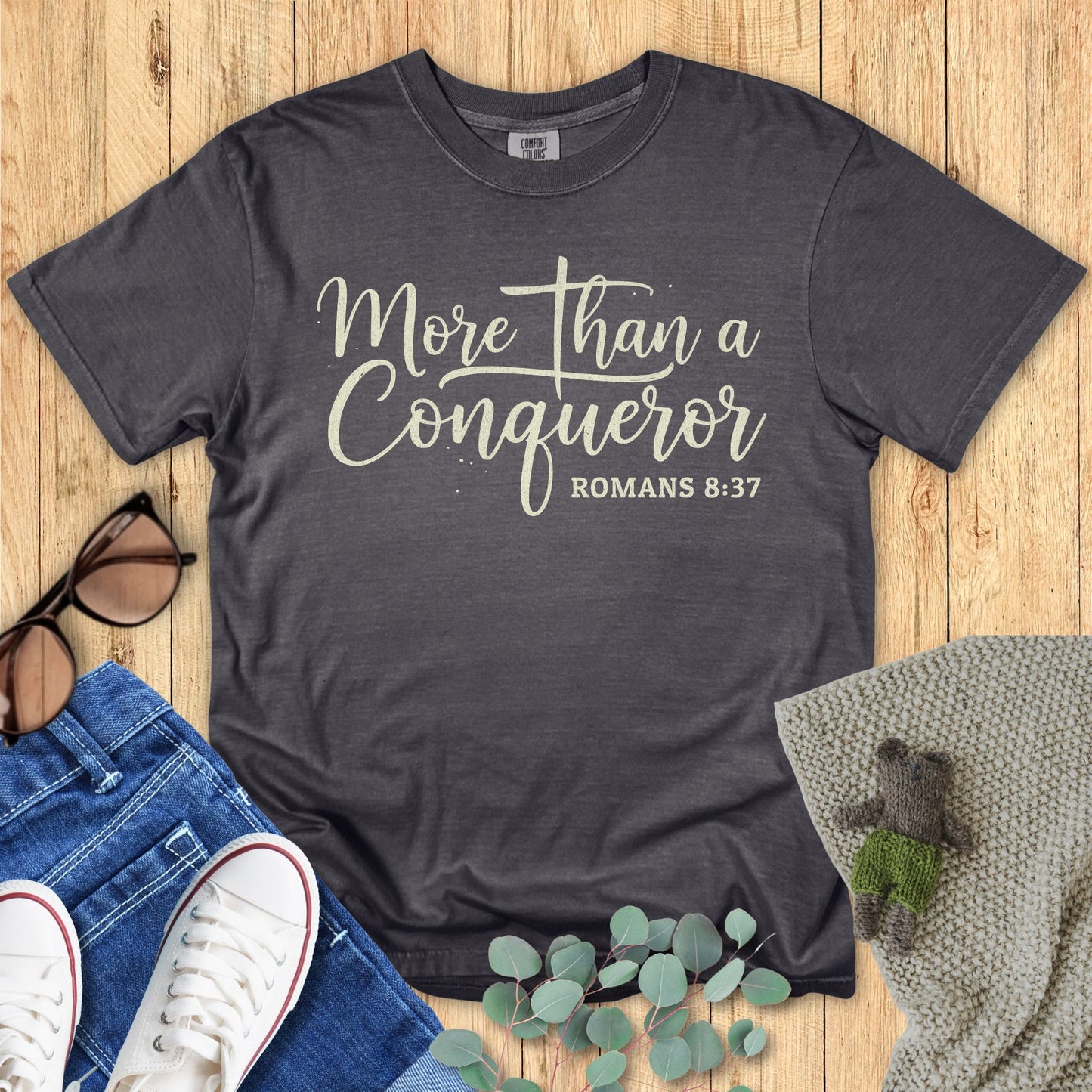 Dark gray t-shirt with 'More than a Conqueror' text on a wooden surface with accessories.