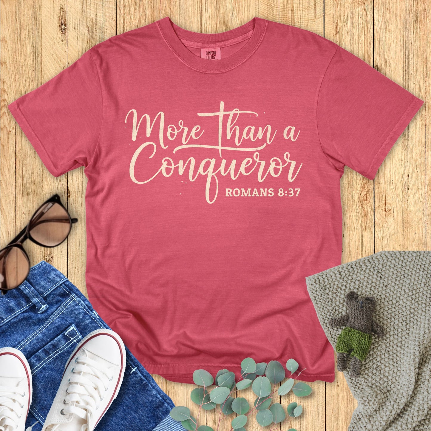 Red t-shirt with 'More than a Conqueror' text on a wooden surface with accessories.