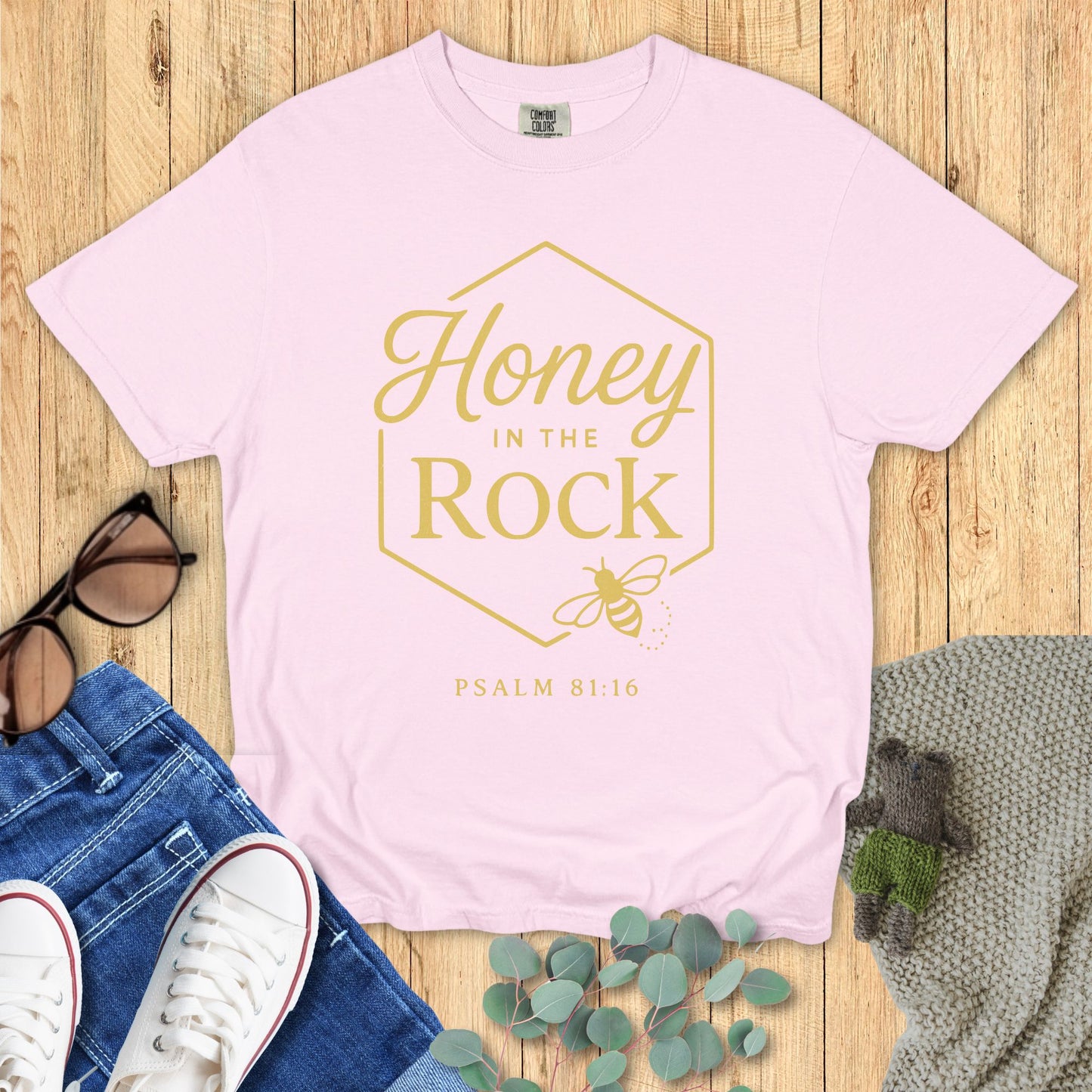 Pink t-shirt with 'Honey in the Rock' design on a wooden surface with jeans, sneakers, and leaves.