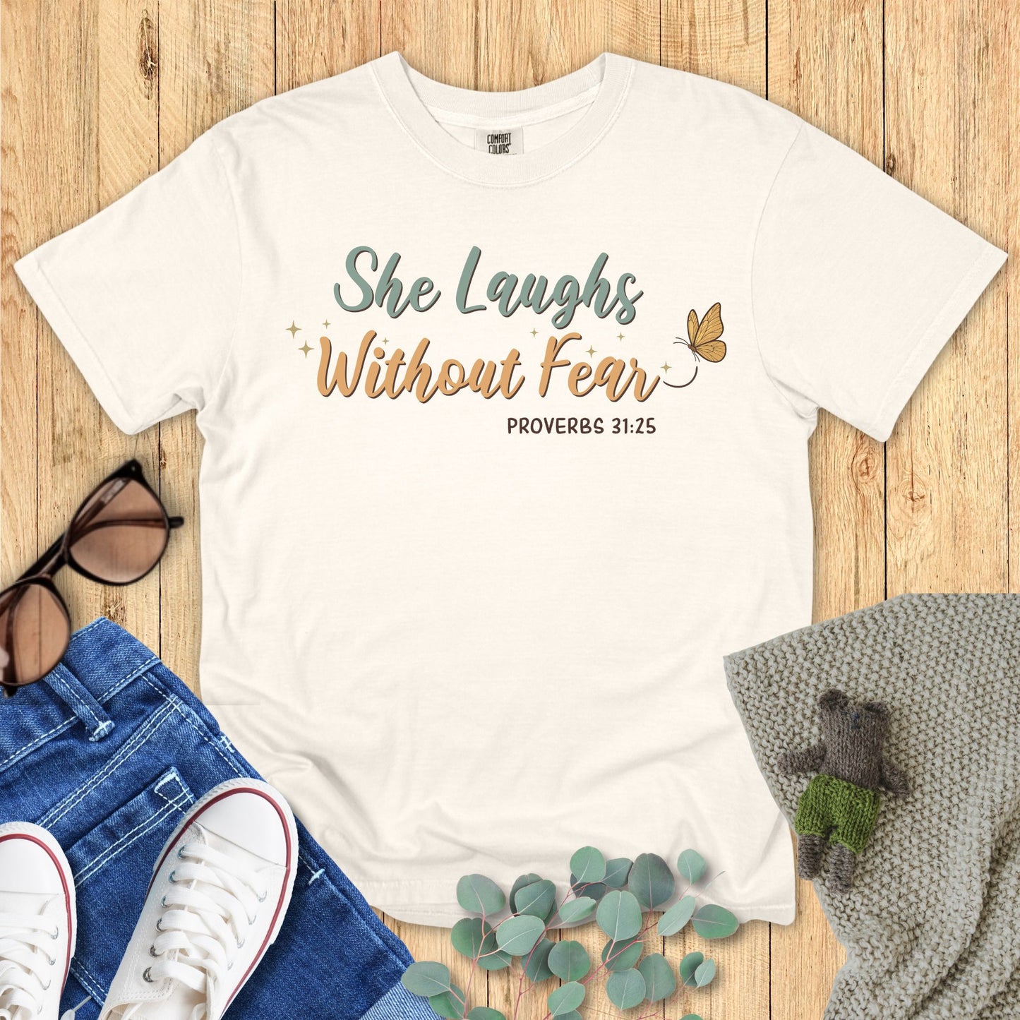 Ivory White t-shirt with 'She Laughs Without Fear' text on a wooden surface with jeans, shoes, and a plant.