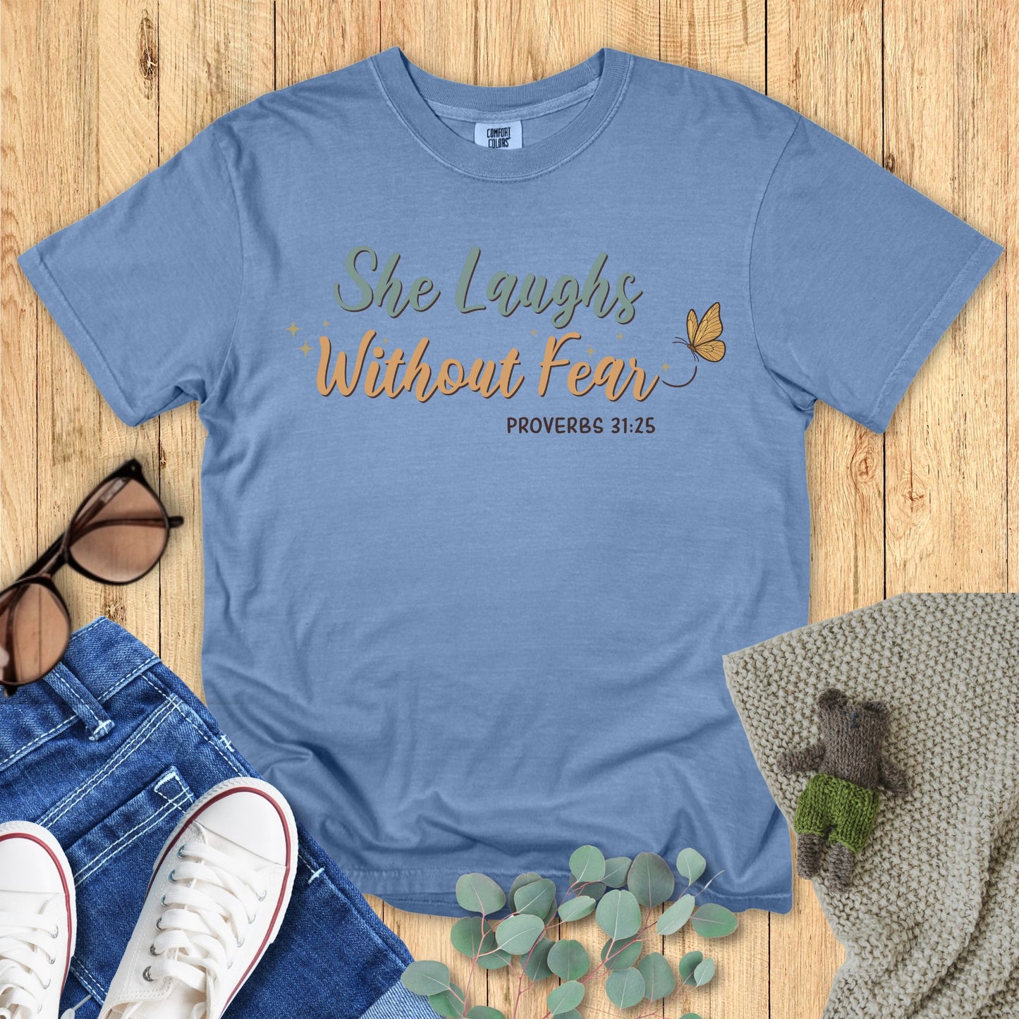 Blue t-shirt with 'She Laughs Without Fear' text on a wooden surface with accessories.