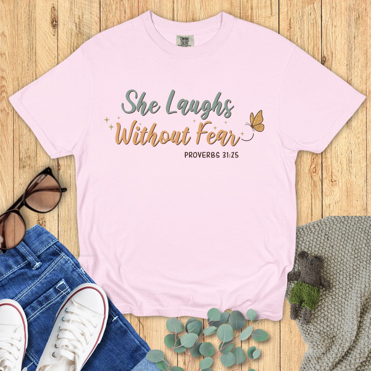Pink t-shirt with 'She Laughs Without Fear' text on a wooden surface with accessories.