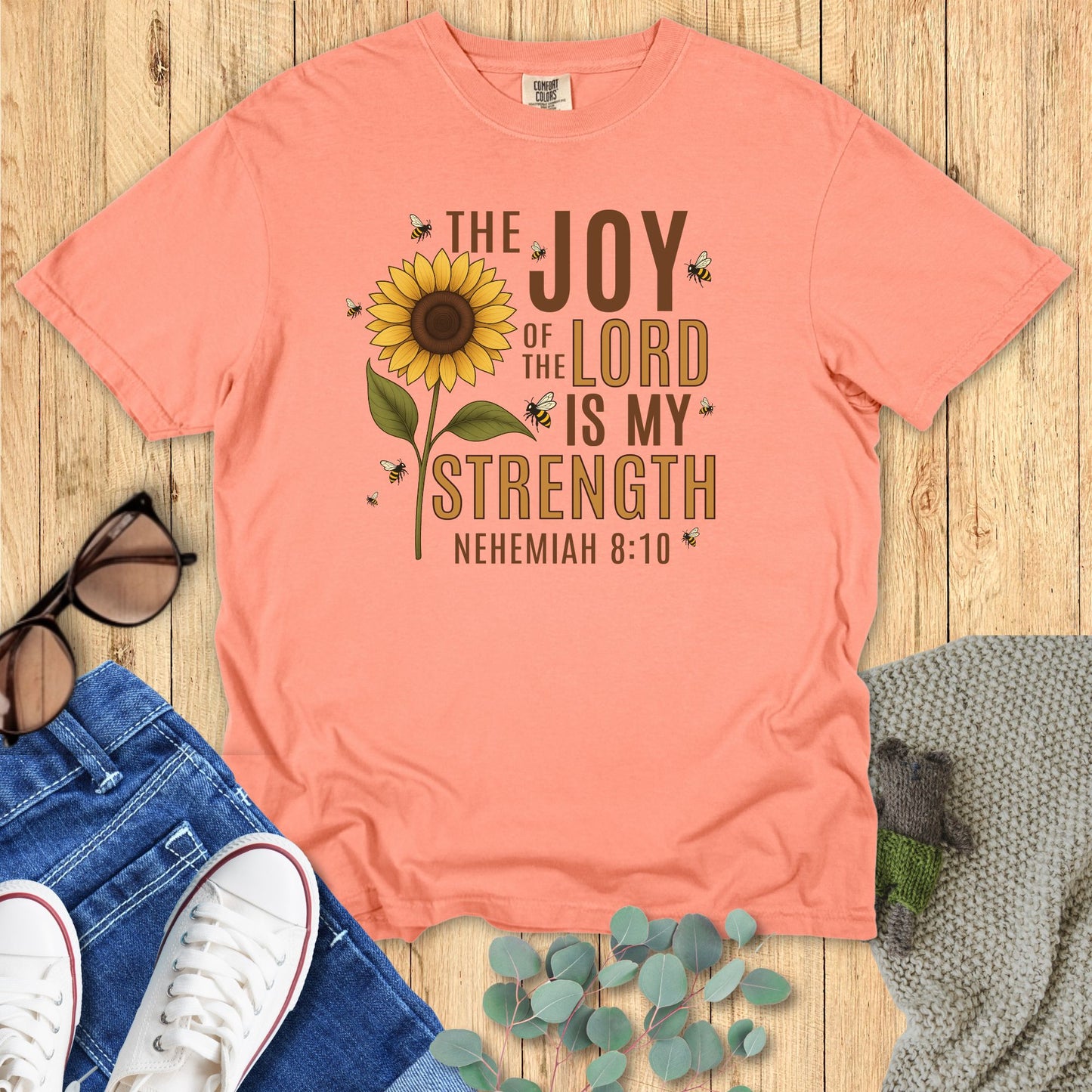Peach t-shirt with sunflower design and text on a wooden surface with accessories.