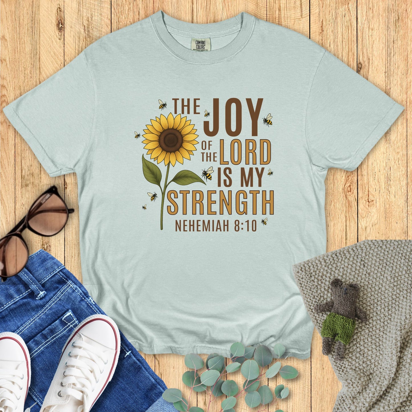 Light blue t-shirt with sunflower graphic and text on a wooden surface with accessories.