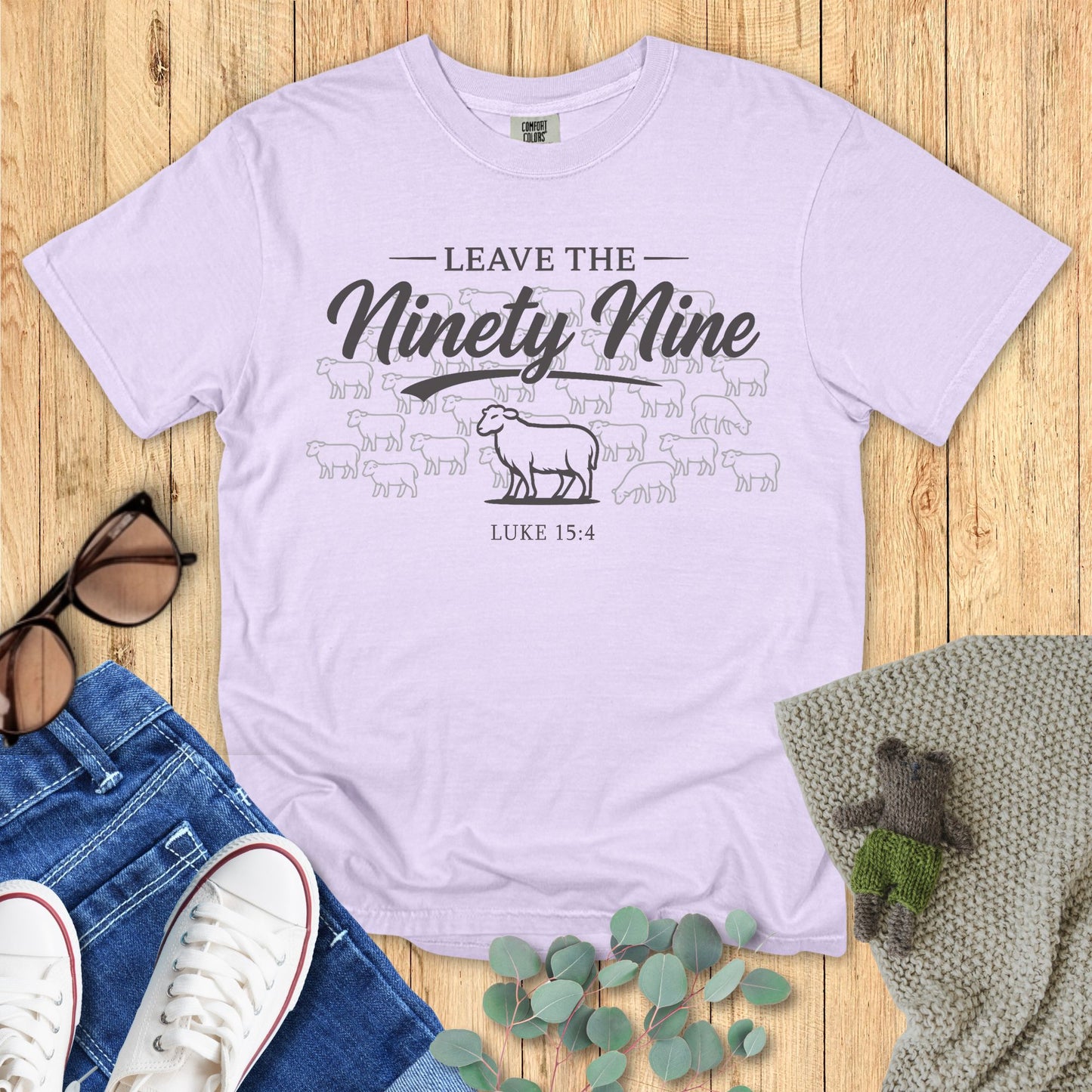 Lavender t-shirt with text and graphics on a wooden surface with accessories.