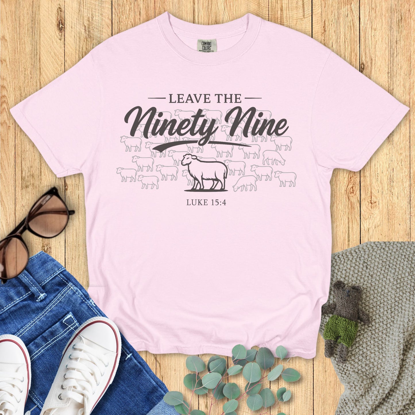 Pink t-shirt with graphic design and text on a wooden surface with jeans and sneakers.