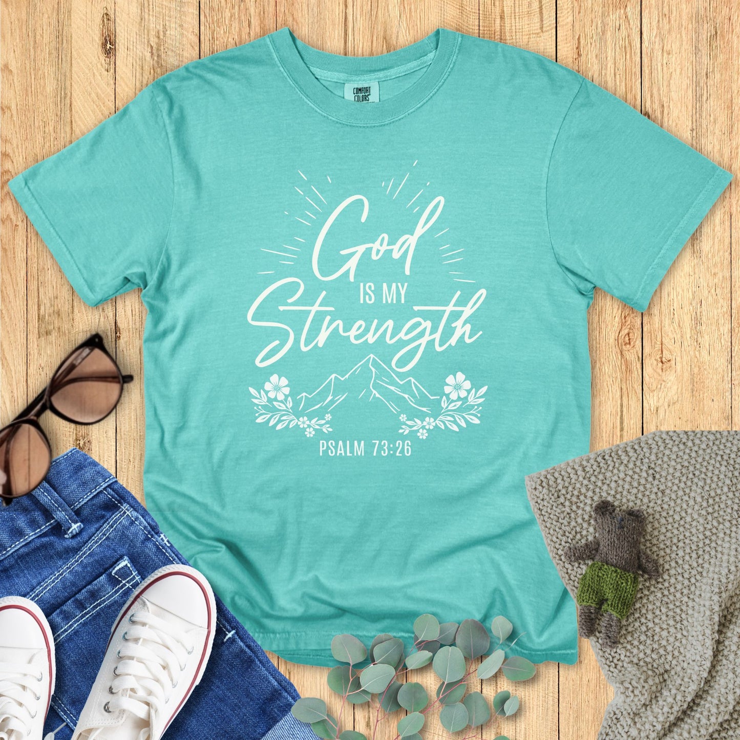 Teal t-shirt with 'God is my strength' text on a wooden surface with accessories.