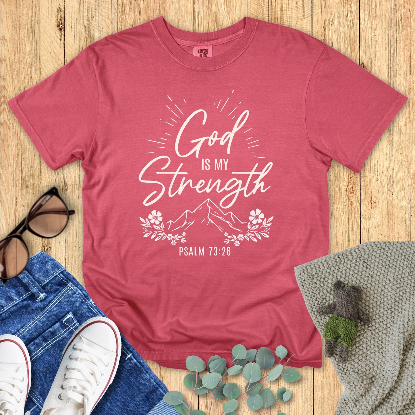 Red t-shirt with 'God is my strength' text on a wooden surface with accessories.