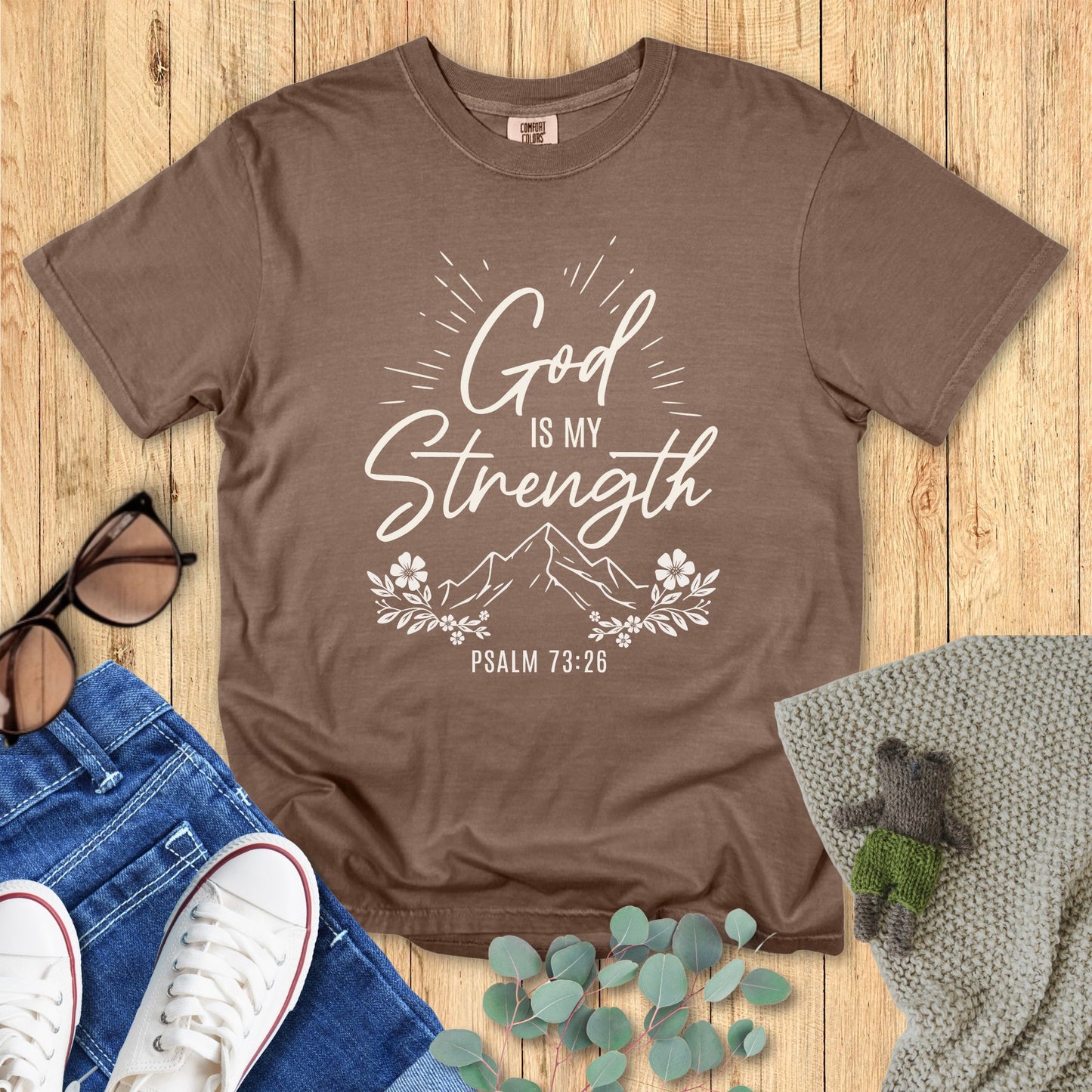 Brown t-shirt with 'God is my strength' text on a wooden surface with accessories.