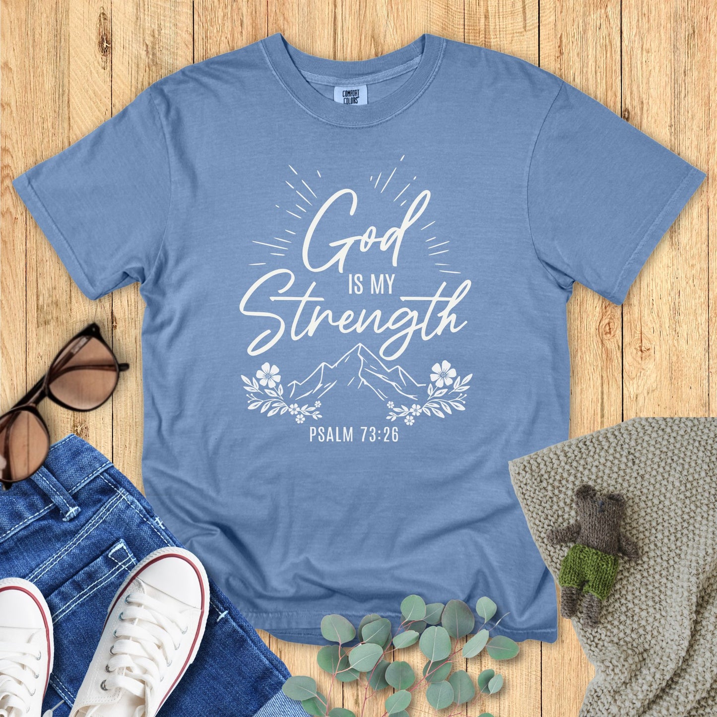 Blue t-shirt with 'God is my strength' text on a wooden surface with accessories.