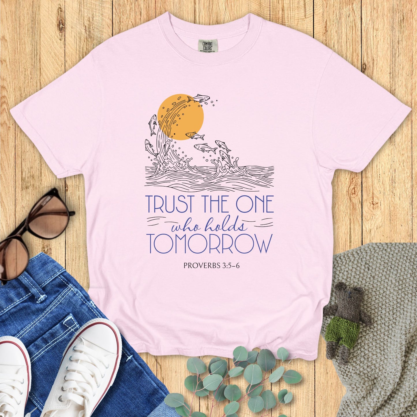 Pink t-shirt with a graphic design and text on a wooden surface with jeans and sneakers.