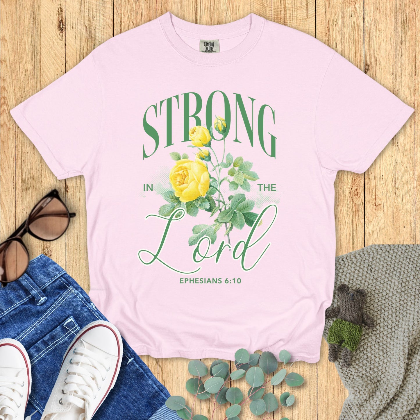Pink t-shirt with 'Strong in the Lord' design on a wooden surface with accessories.