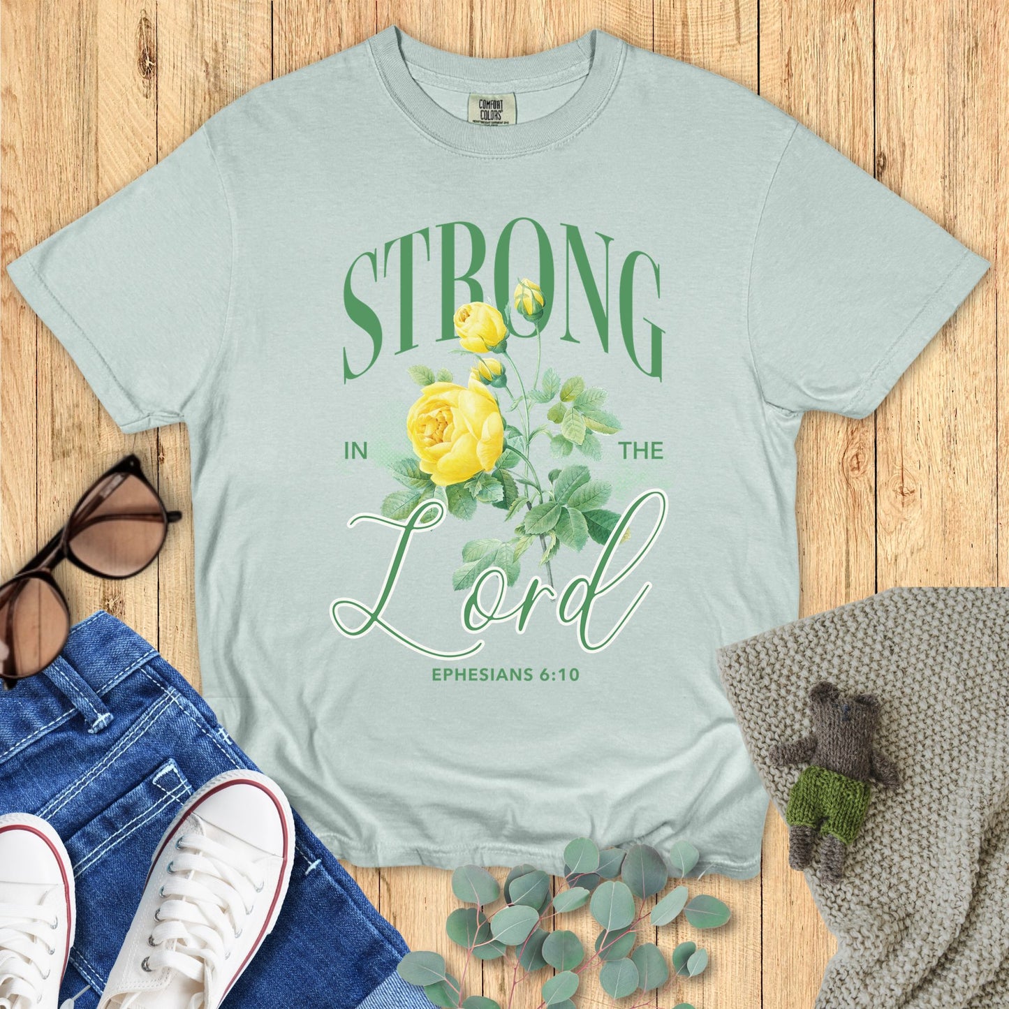 Light green t-shirt with 'Strong in the Lord' design on a wooden surface with accessories.