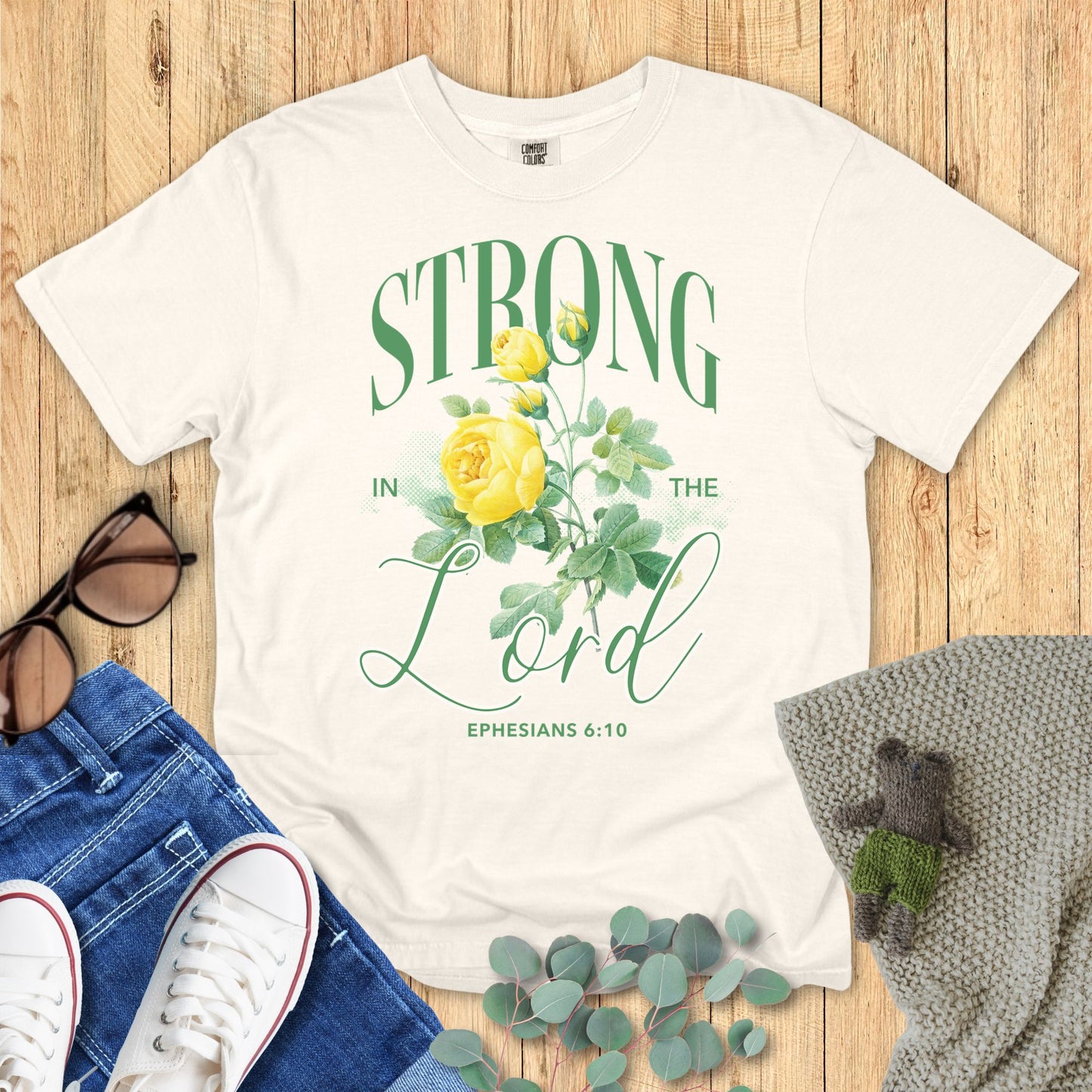 Ivory t-shirt with 'Strong in the Lord' design on a wooden surface with accessories.