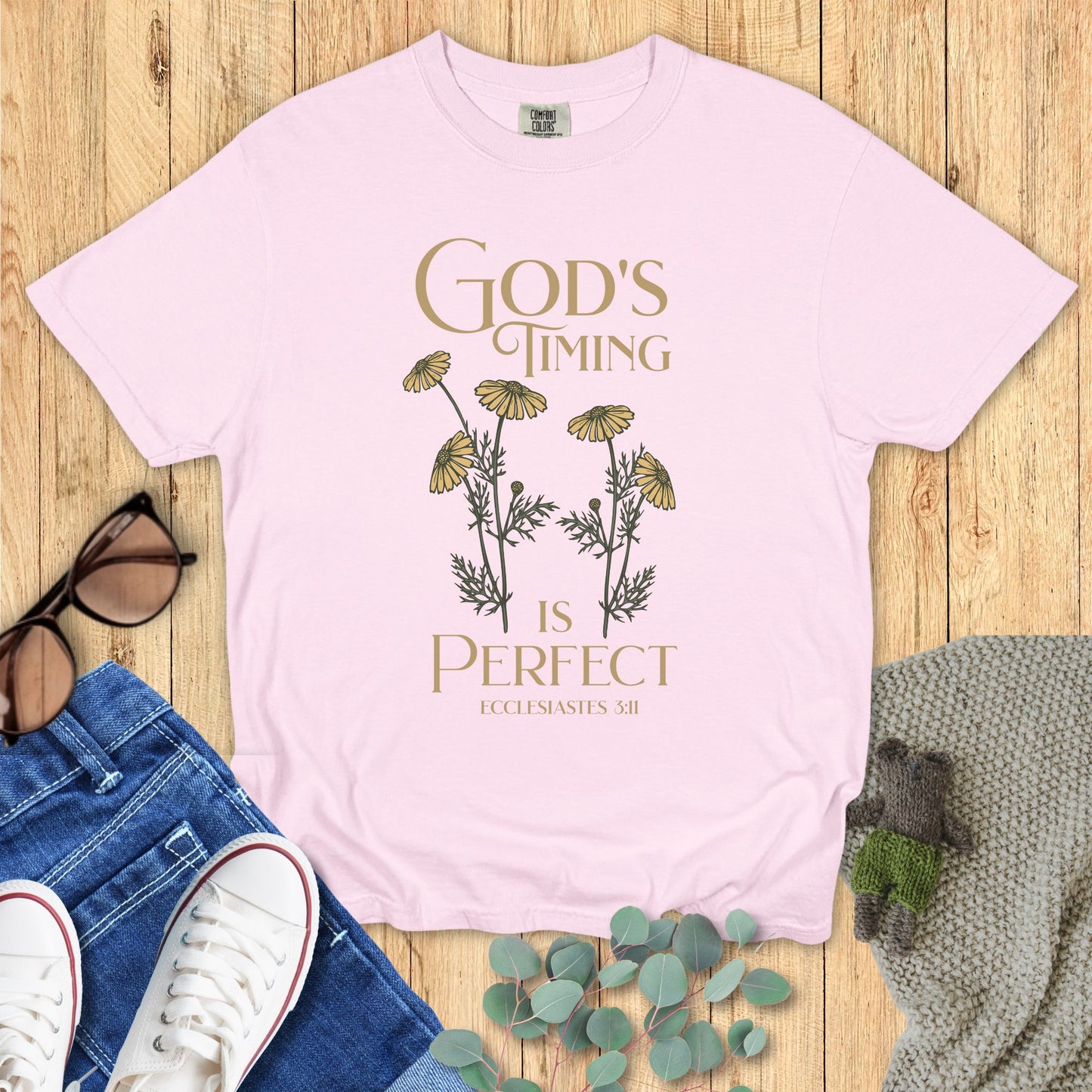 Pink t-shirt with 'God's Timing is Perfect' text and floral design on a wooden surface with jeans and sneakers.