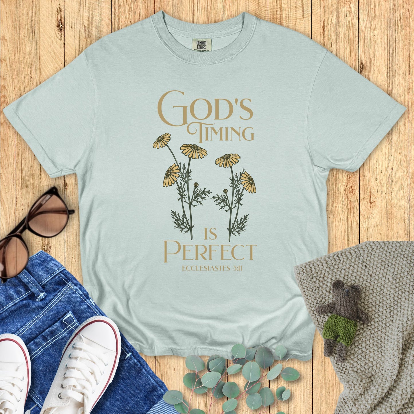 Light blue t-shirt with floral design and text on a wooden surface with accessories.