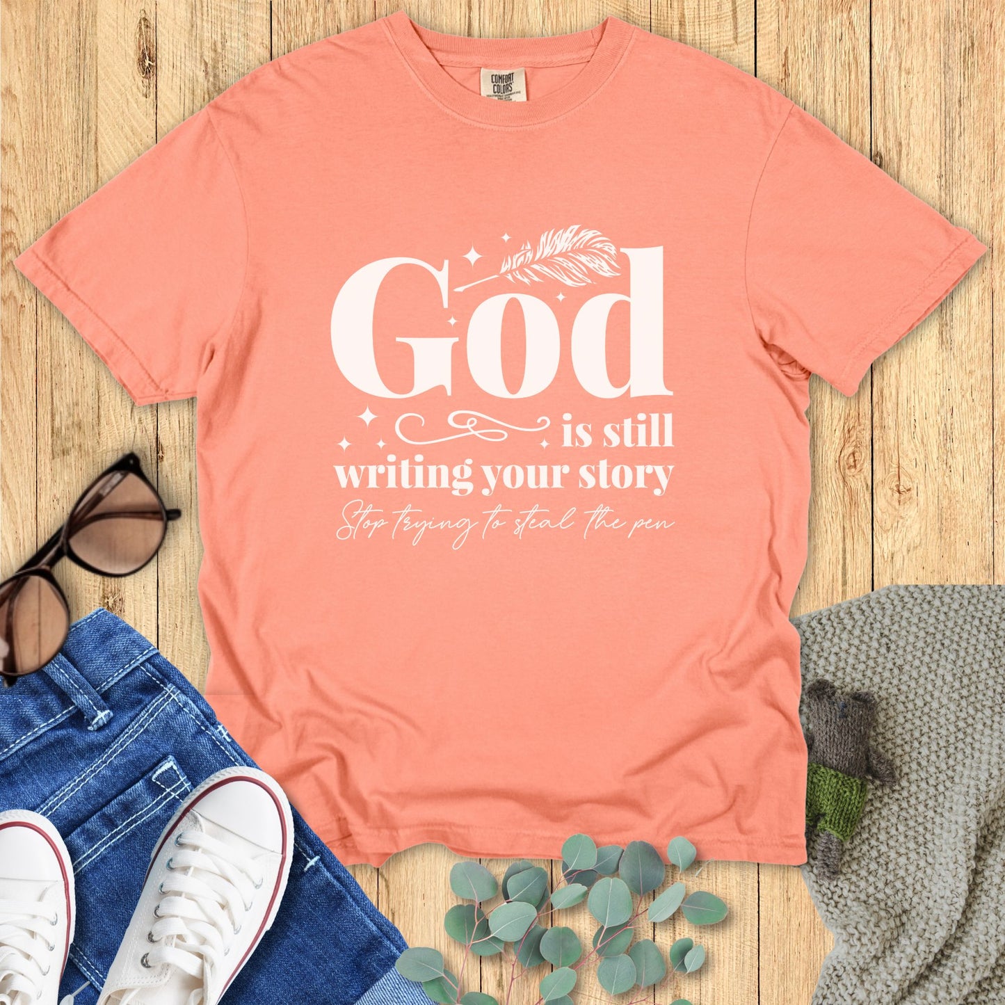 Coral t-shirt with motivational quote on a wooden surface with accessories.