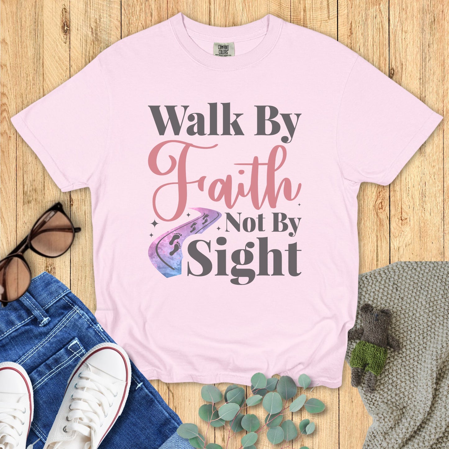 Pink t-shirt with 'Walk By Faith Not By Sight' text on a wooden surface with jeans and sneakers.