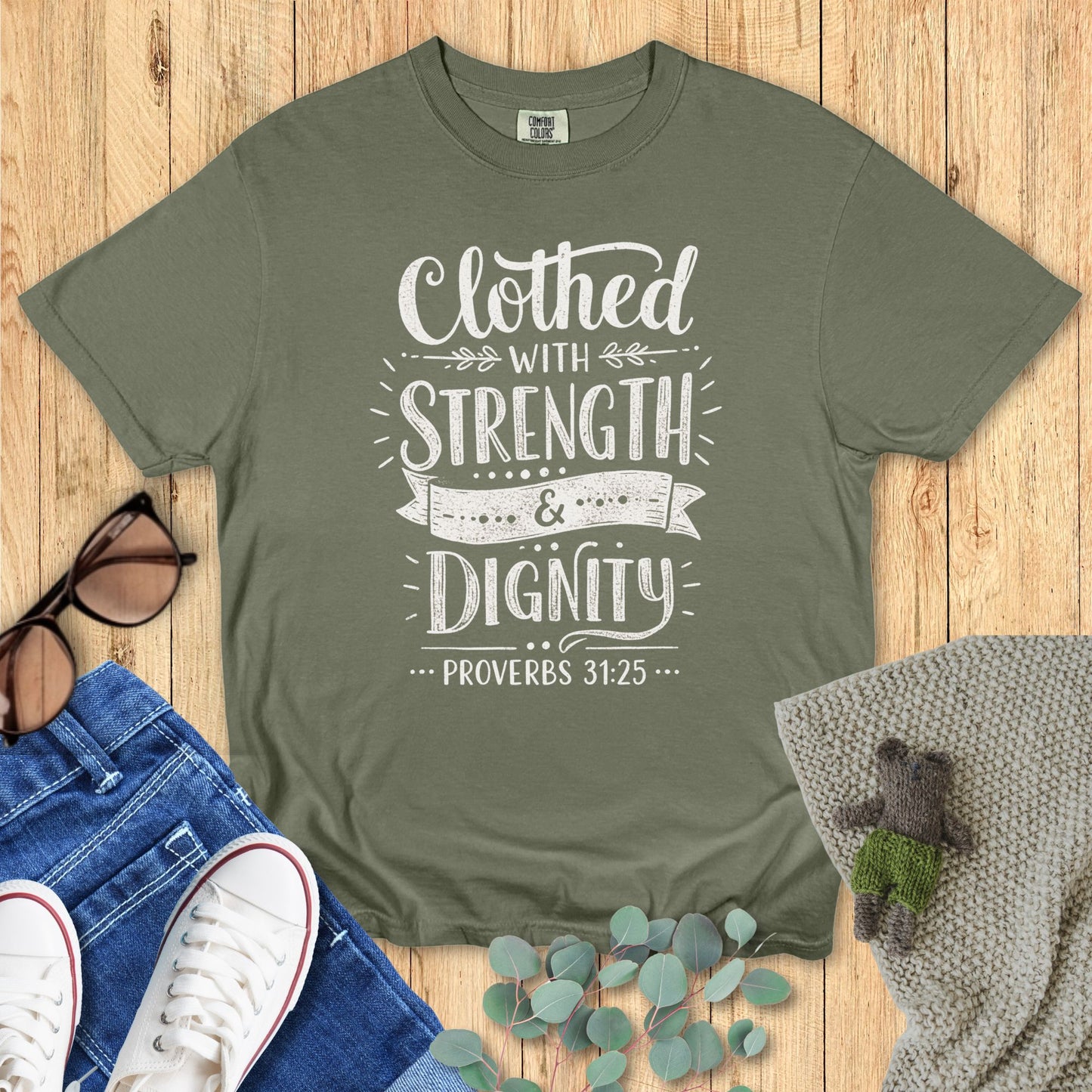 Green t-shirt with 'Clothed with Strength & Dignity' text on a wooden surface with accessories.